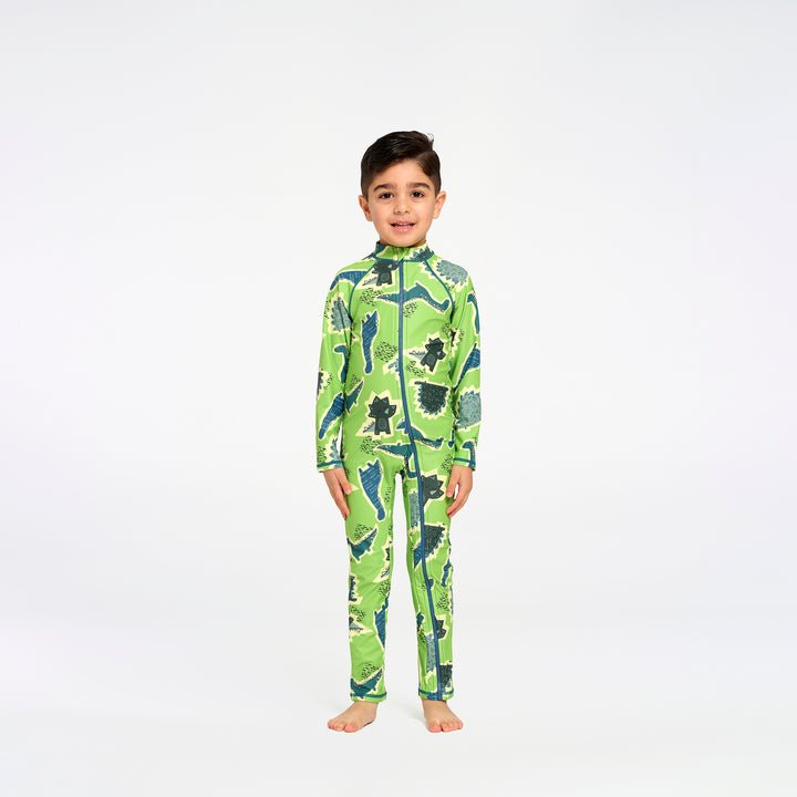 A young boy stands barefoot against a plain white studio background, wearing a long-sleeve, high-collar one-piece swimsuit with an allover dinosaur print in bright green, dark green, and blue.