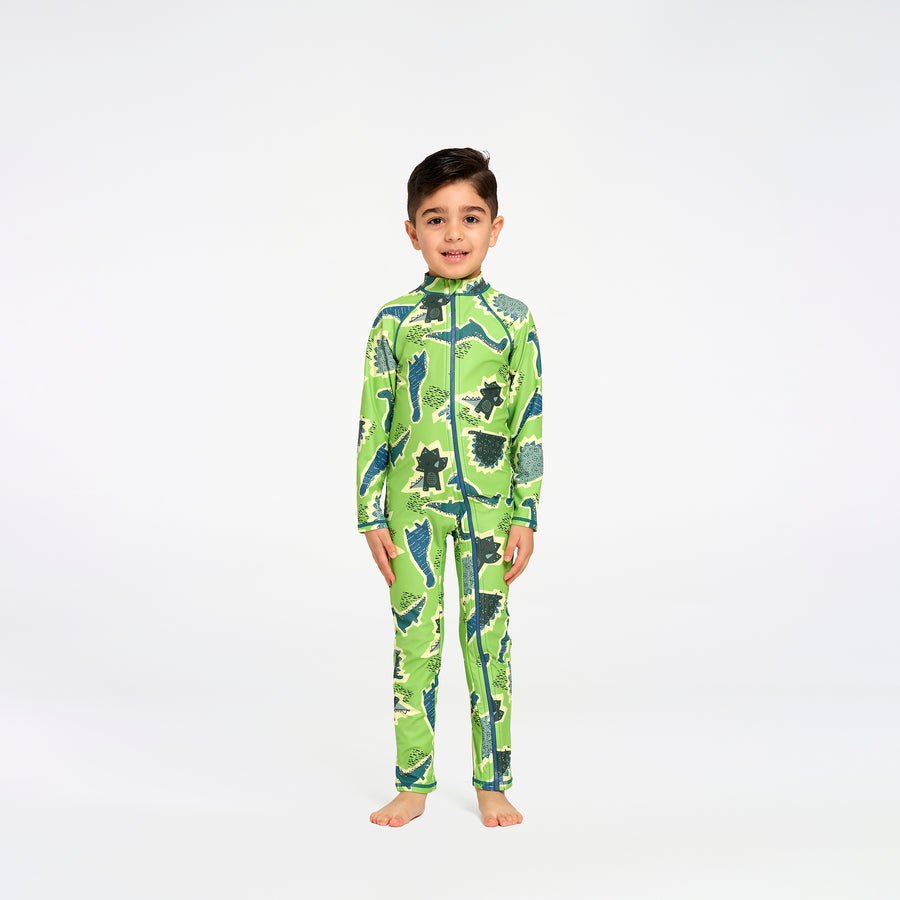 A young boy stands barefoot against a plain white studio background, wearing a long-sleeve, high-collar one-piece swimsuit with an allover dinosaur print in bright green, dark green, and blue.