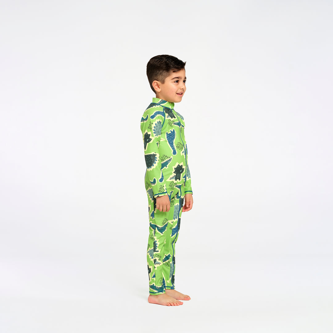 A young boy standing in profile wearing a green long-sleeve swim suit with a dinosaur pattern, photographed against a seamless white studio background.