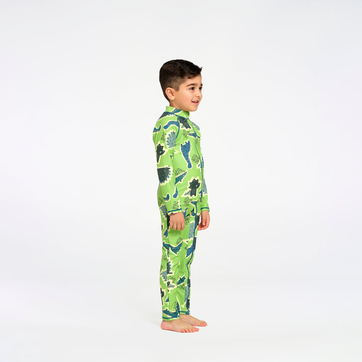 A young boy standing in profile wearing a green long-sleeve swim suit with a dinosaur pattern, photographed against a seamless white studio background.