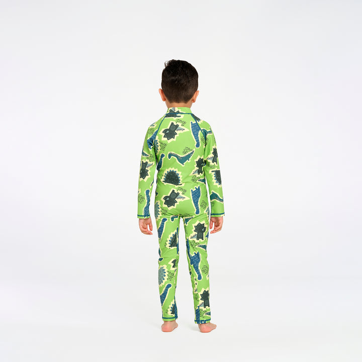 A young boy viewed from behind wearing a green long-sleeve swimsuit with a dinosaur print, standing against a plain white studio background.