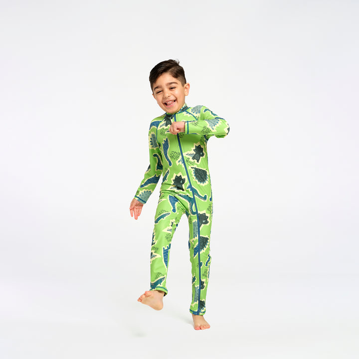 A young boy wearing a green full-zip one-piece swimsuit with a dinosaur pattern, posing and smiling against a solid white studio background.