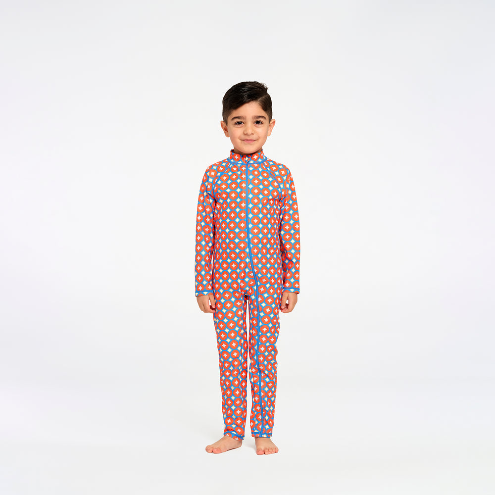 A young boy stands barefoot against a plain white studio background, wearing a long-sleeve one-piece swimsuit with an orange base and a repeating pattern of blue and yellow dots, outlined with blue piping.