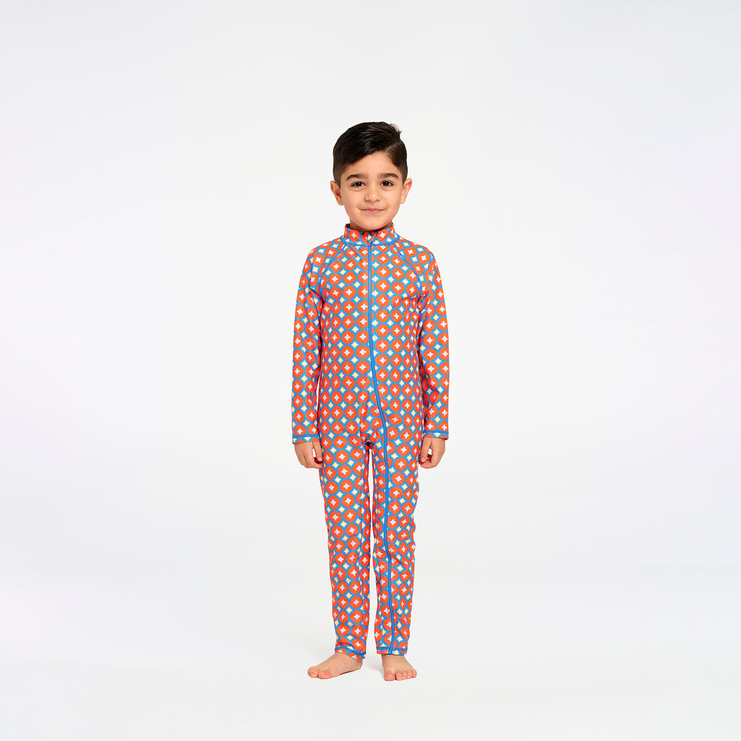 A young boy stands barefoot against a plain white studio background, wearing a long-sleeve one-piece swimsuit with an orange base and a repeating pattern of blue and yellow dots, outlined with blue piping.