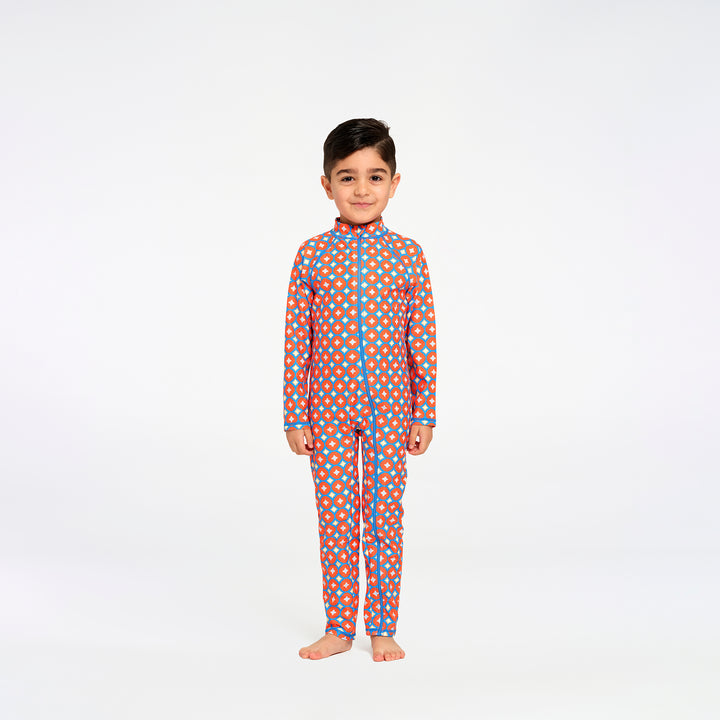 A young boy stands barefoot against a plain white studio background, wearing a long-sleeve one-piece swimsuit with an orange base and a repeating pattern of blue and yellow dots, outlined with blue piping.