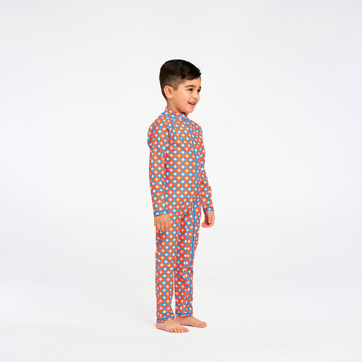 A young boy standing in profile wearing a long-sleeve, full-length one-piece swimsuit with an orange, blue, and white geometric pattern, set against a plain white studio background.