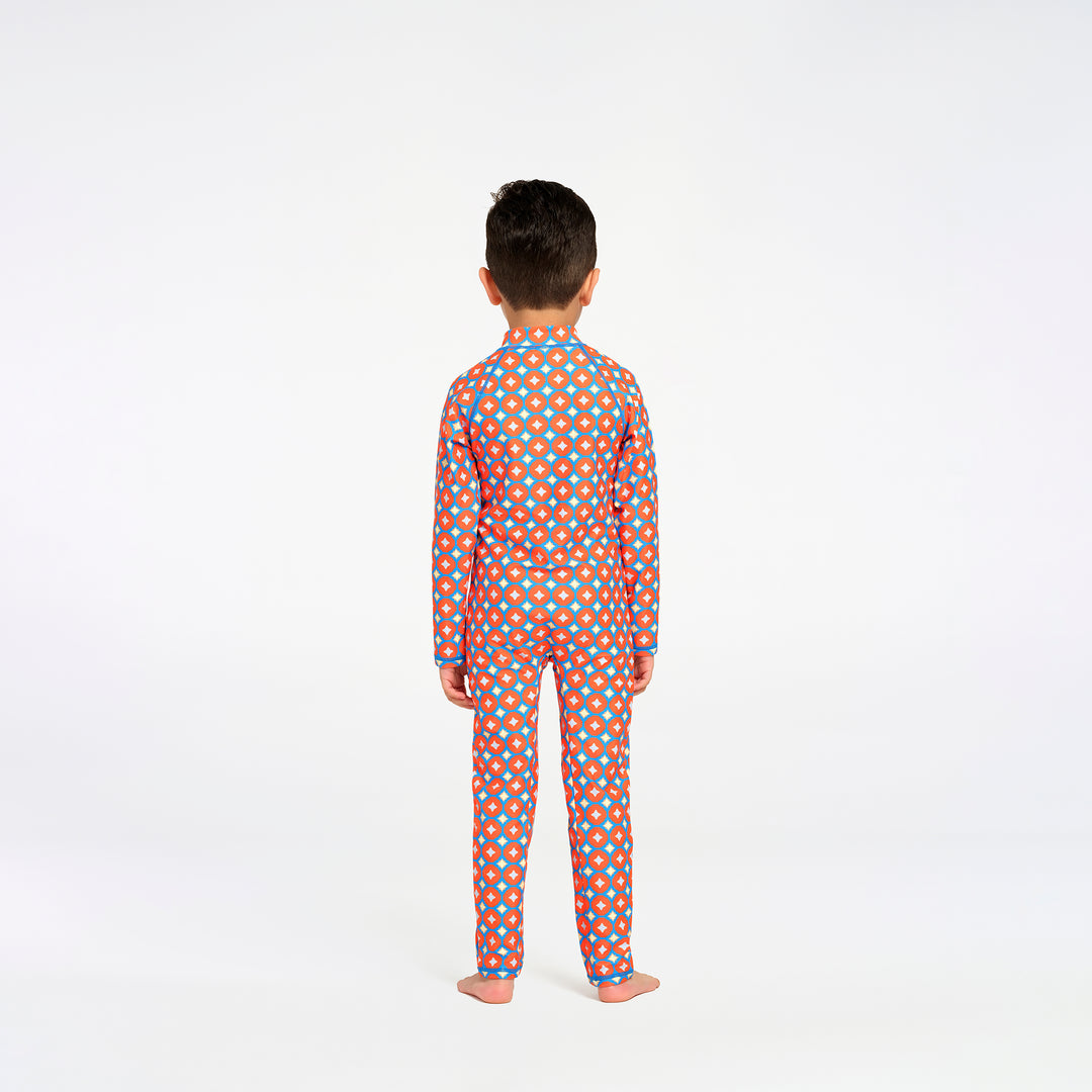 A child standing with their back to the camera, wearing a long-sleeve swimsuit with an orange and blue polka dot pattern on a white studio background.