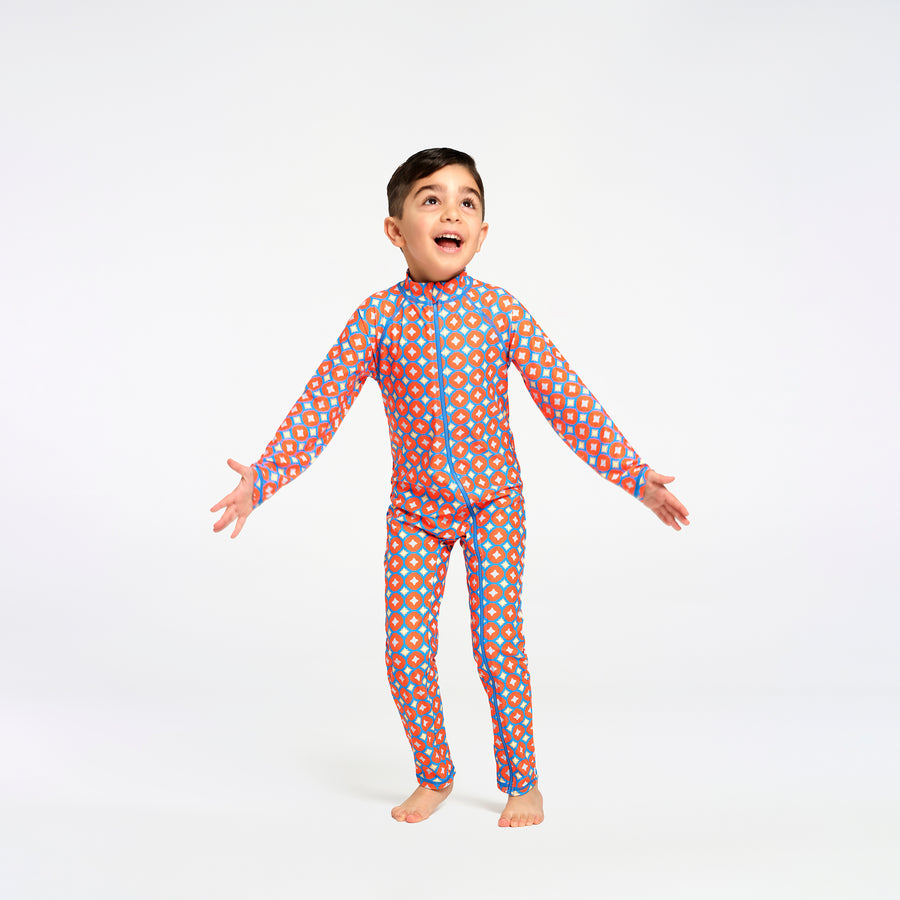 A young boy stands barefoot against a white studio background, wearing a full-length one-piece zip-up swimsuit with a bright orange base and an allover pattern of blue and yellow circles.