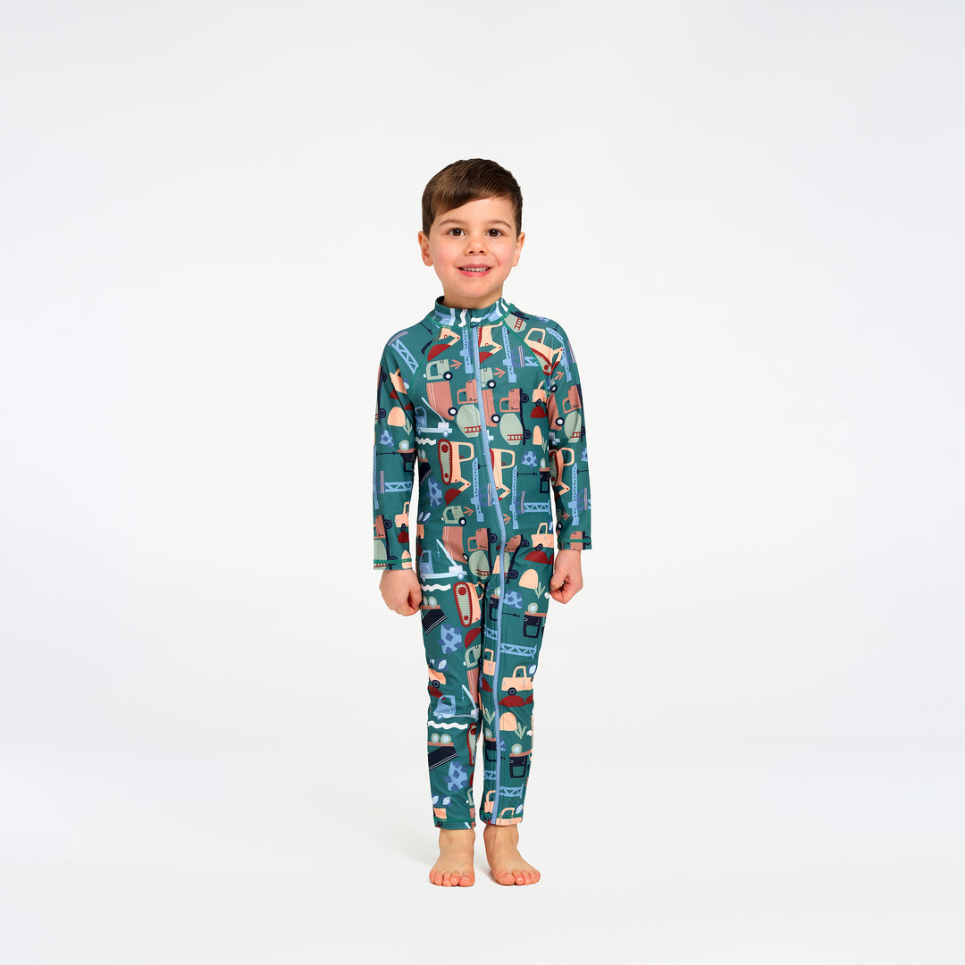A young boy stands barefoot against a plain white studio background, wearing a long-sleeve, full-length zip-up one-piece swimsuit with an allover transport print in teal, blue, beige, and brown tones.