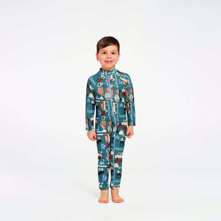A young boy stands barefoot against a plain white studio background, wearing a long-sleeve, full-length zip-up one-piece swimsuit with an allover transport print in teal, blue, beige, and brown tones.