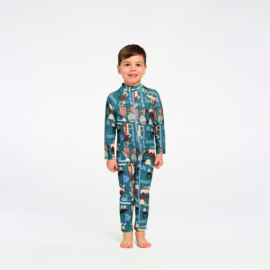 A young boy stands barefoot against a plain white studio background, wearing a long-sleeve, full-length zip-up one-piece swimsuit with an allover transport print in teal, blue, beige, and brown tones.