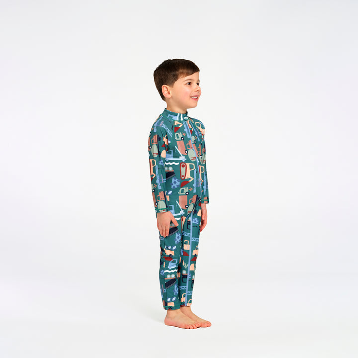 A young boy stands in profile wearing a one-piece long-sleeve swimsuit with a colorful cartoon pattern on a teal background, photographed against a seamless white studio background.