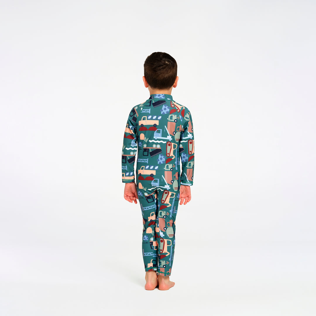 A child standing with their back to the camera, wearing a long-sleeve swimsuit with a colorful cartoon truck pattern on a teal background, set against a plain white studio background.