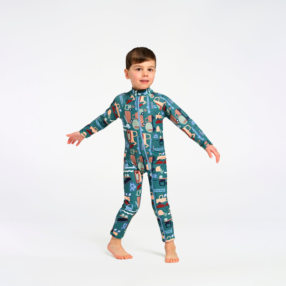 A young boy wearing a full-body teal and multicolor patterned swimsuit, standing against a plain white studio background.