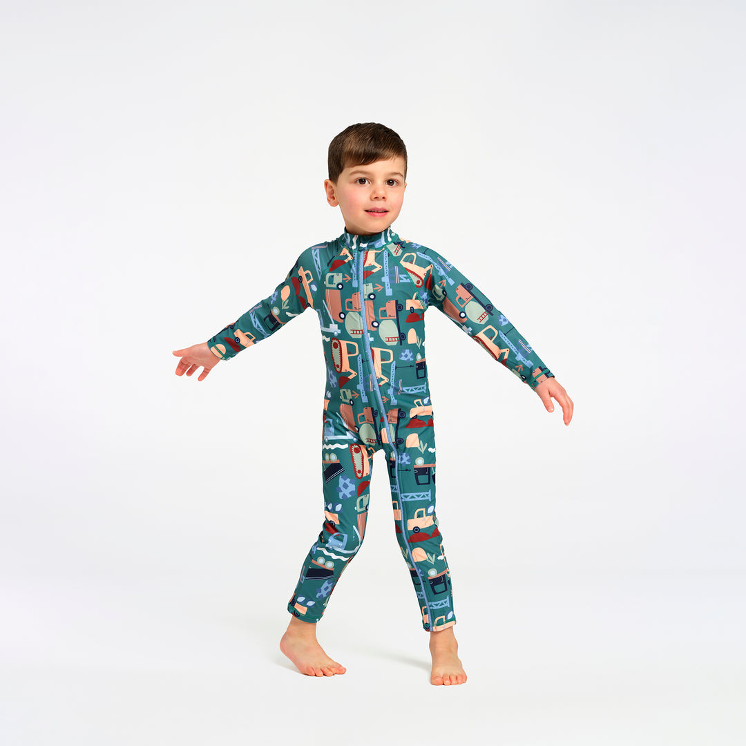A young boy wearing a full-body teal and multicolor patterned swimsuit, standing against a plain white studio background.