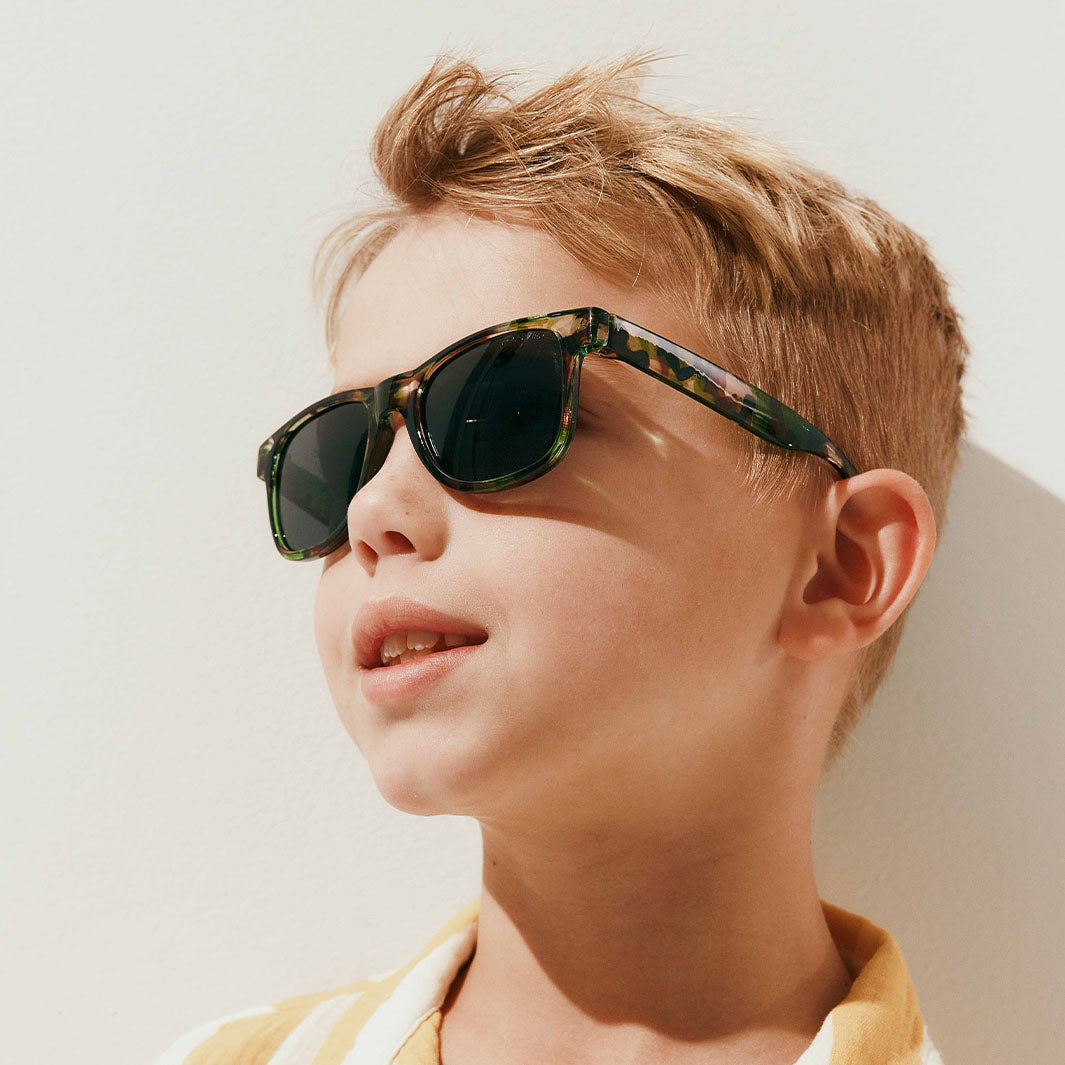 A close-up of a young boy with blond hair wearing tortoiseshell sunglasses with dark lenses, set against a bright, sunlit off-white background.