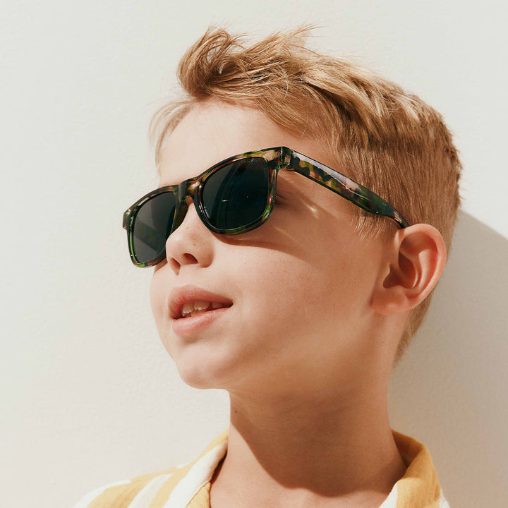 A close-up of a young boy with blond hair wearing tortoiseshell sunglasses with dark lenses, set against a bright, sunlit off-white background.