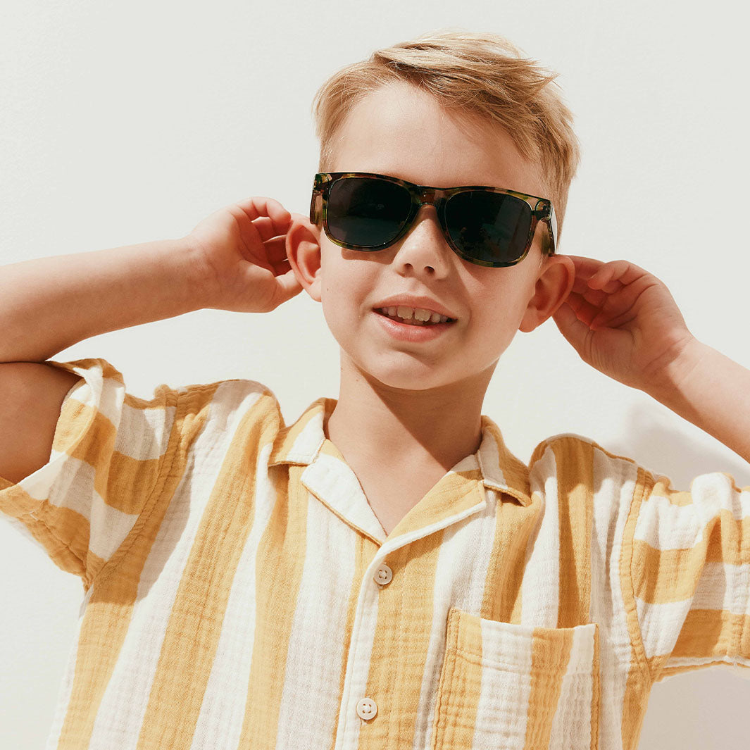 A young boy wearing dark tortoiseshell sunglasses and a yellow and white striped short-sleeve shirt, posing against a clean white background.