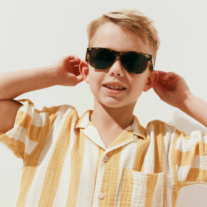 A young boy wearing dark tortoiseshell sunglasses and a yellow and white striped short-sleeve shirt, posing against a clean white background.