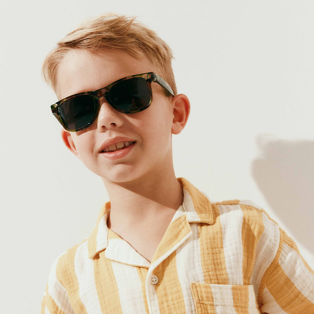A young boy with blond hair wearing dark tortoiseshell sunglasses and a yellow-and-white striped shirt, photographed against a clean white background with a soft shadow.