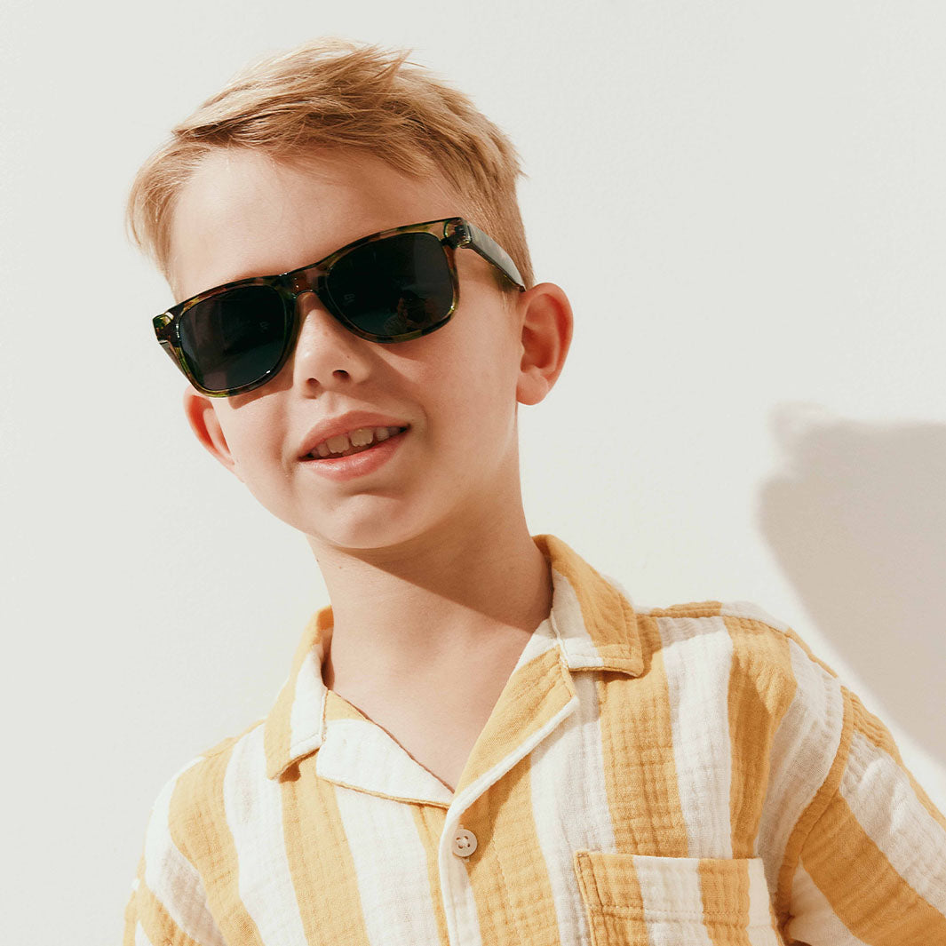 A young boy with blond hair wearing dark tortoiseshell sunglasses and a yellow-and-white striped shirt, photographed against a clean white background with a soft shadow.