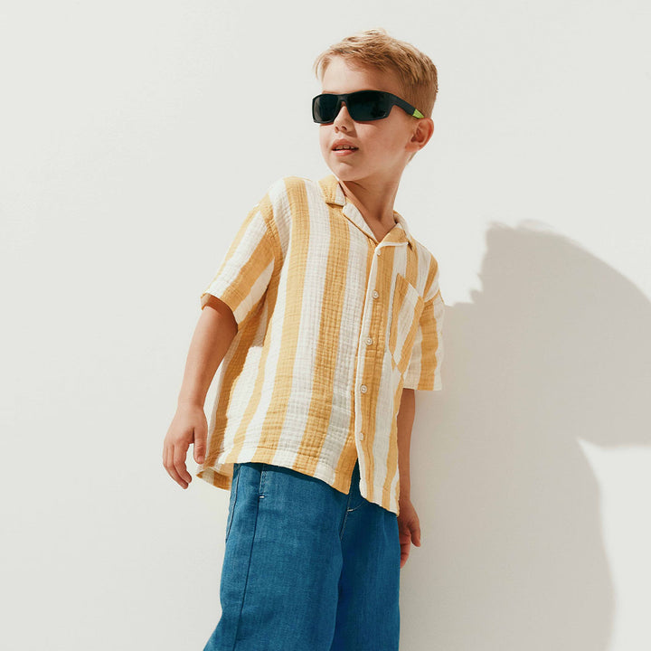 A young boy wearing dark green-tinted sunglasses with a light green temple arm and a yellow-and-white striped shirt