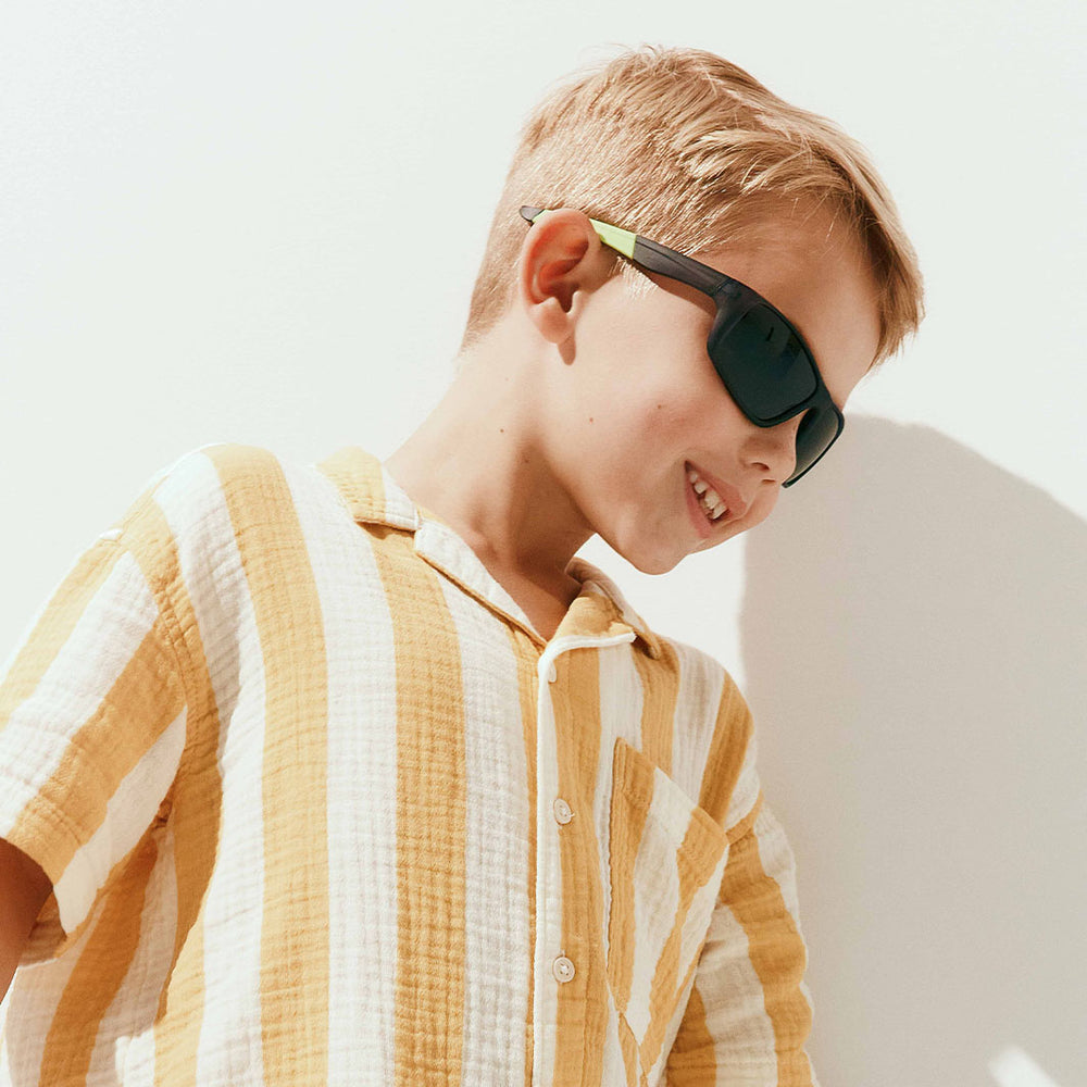 A young boy wearing dark green-tinted sunglasses with a light green temple arm and a yellow-and-white striped shirt, photographed against a bright white wall.
