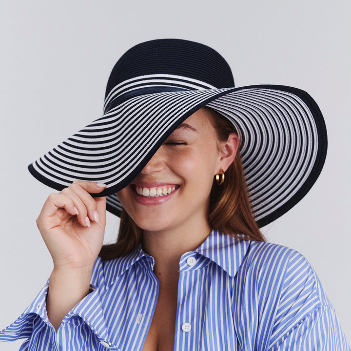 Female model wearing Amy Striped Capeline Hat in Navy/White colour, angled showing wide brim pulled down playfully over face