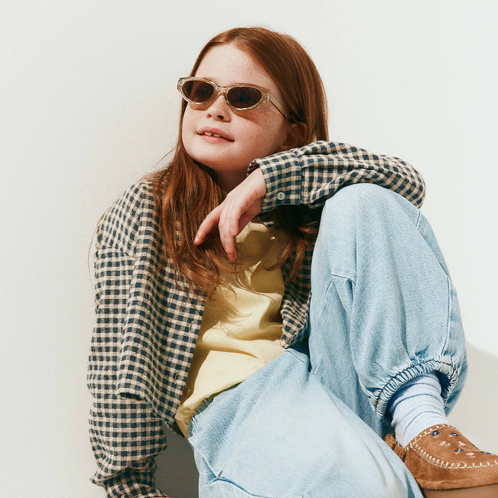 A young girl with red hair wearing round sunglasses, a black and white gingham overshirt over a pale yellow top, light blue baggy jeans, white socks, and brown moccasin shoes, sitting against a plain off-white wall.