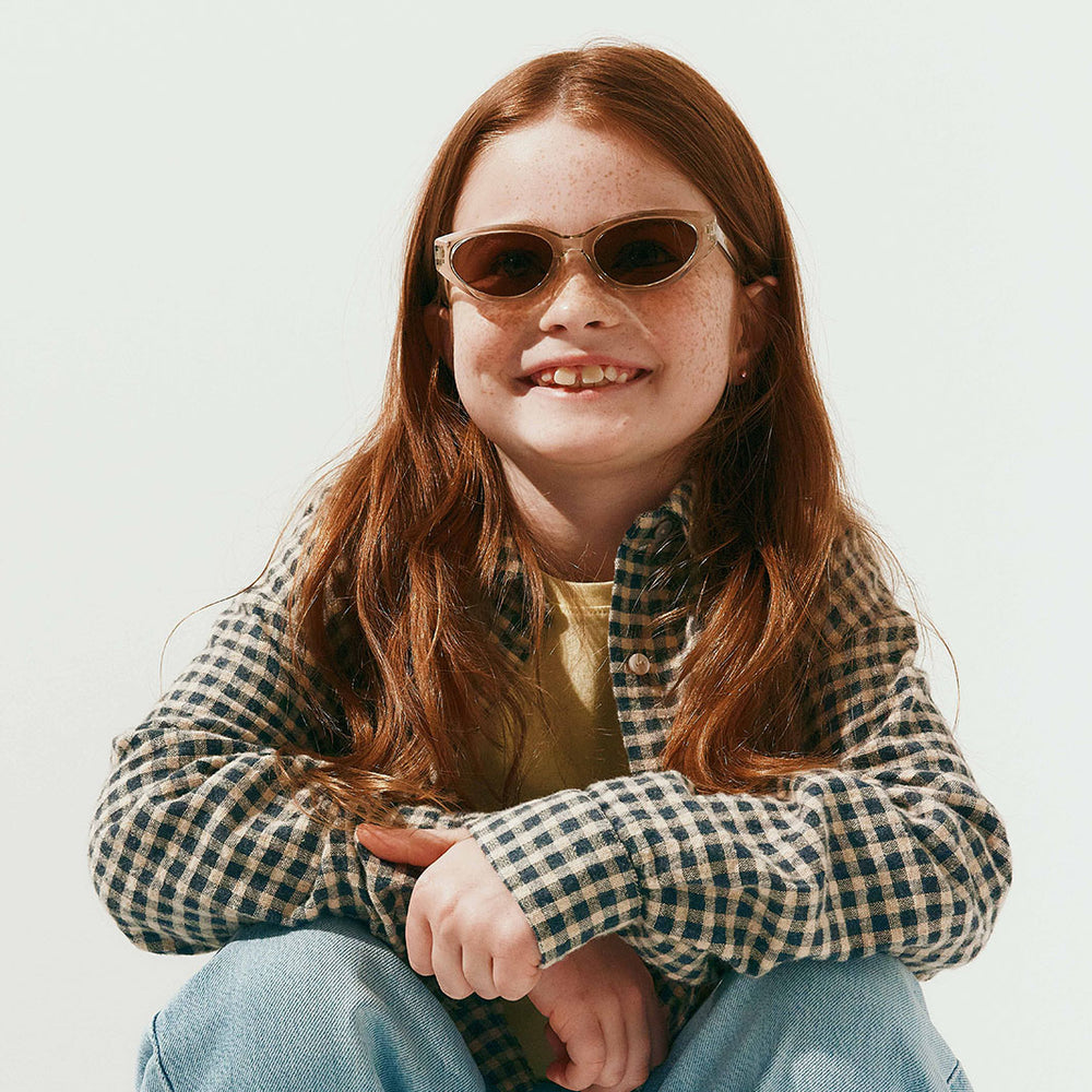 A young girl with long red hair and freckles wearing stylish cat-eye sunglasses, a green and white checkered flannel shirt, and blue jeans, sitting against a solid off-white background.