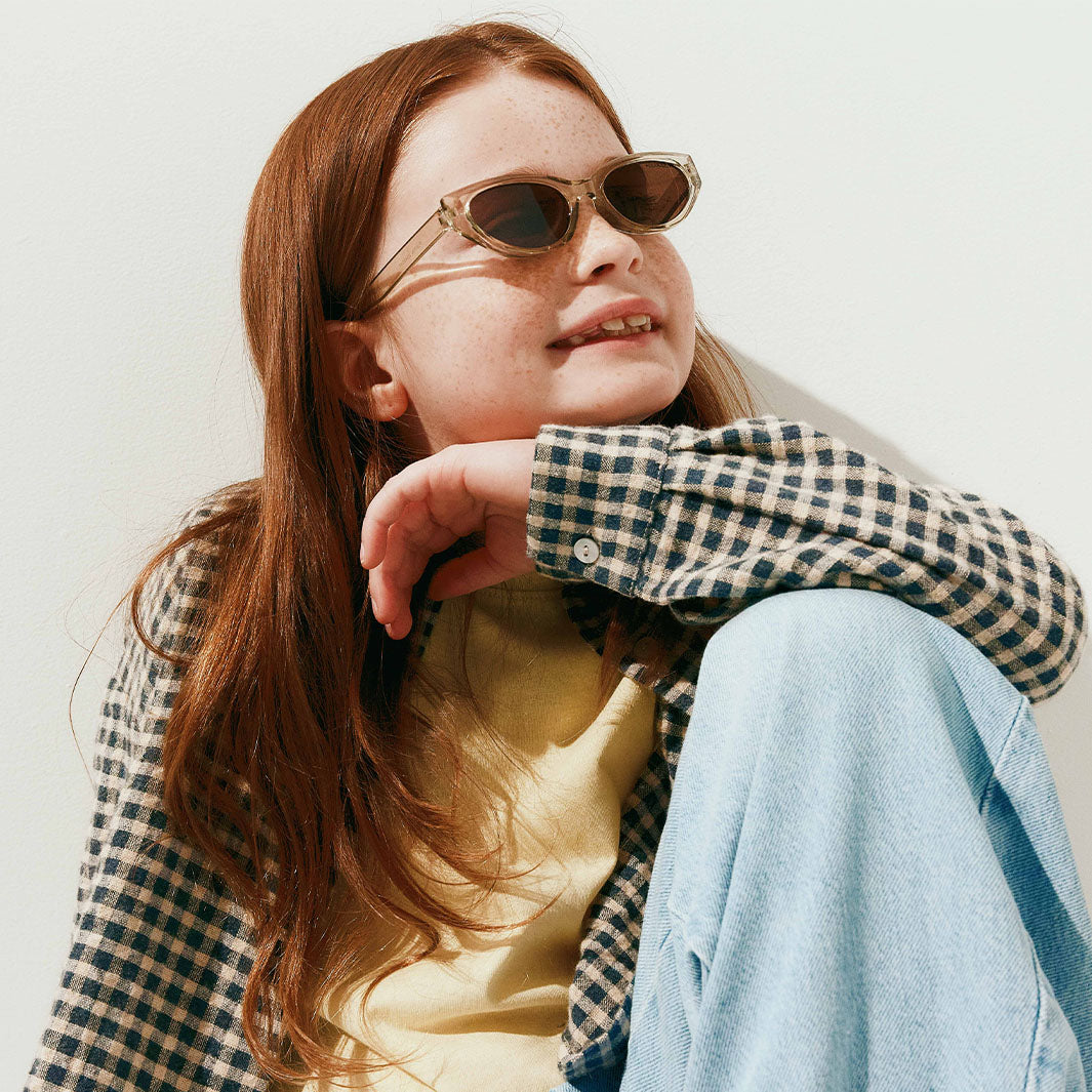 A young girl with long red hair and freckles wearing stylish oval sunglasses with a gold-toned frame and dark lenses, styled with a black and white gingham shirt and light blue jeans against a plain white wall.