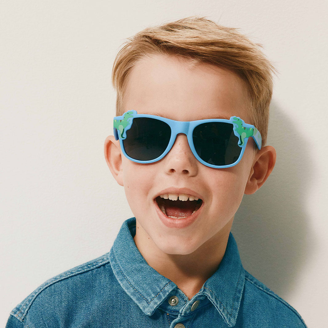 A young boy with blond hair wearing blue dinosaur-shaped sunglasses and a denim shirt, photographed against a plain light background.
