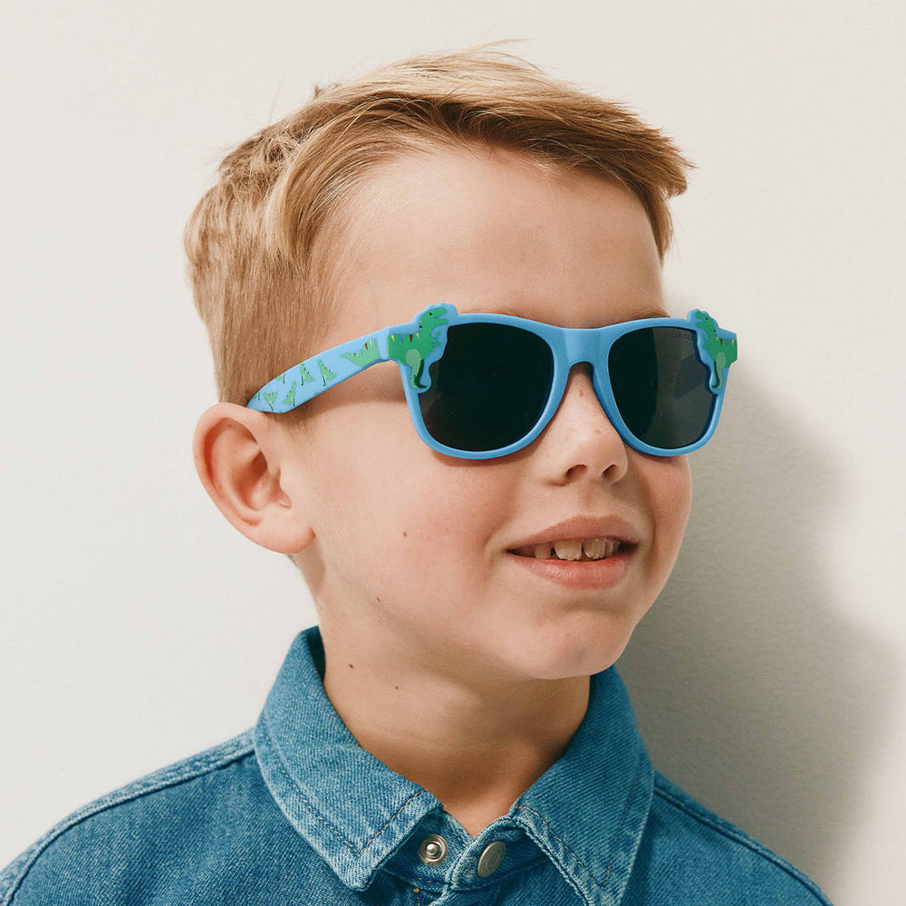 A young boy with blond hair wearing blue sunglasses with a green dinosaur pattern on the arms and a blue denim shirt, photographed against a plain light background.