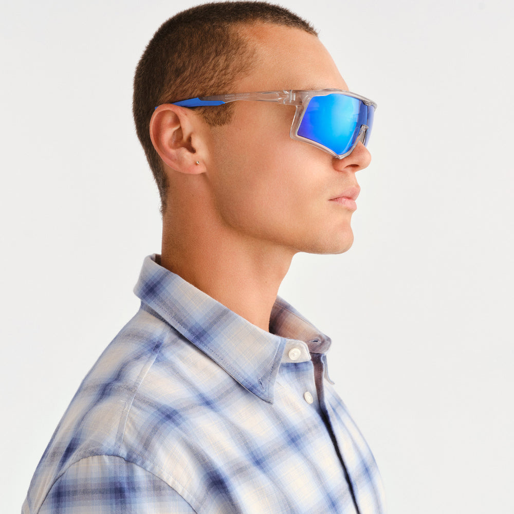 A man in profile wearing reflective blue mirrored shield-style sunglasses with clear frames, paired with a light blue and white plaid button-down shirt, against a plain light gray background.