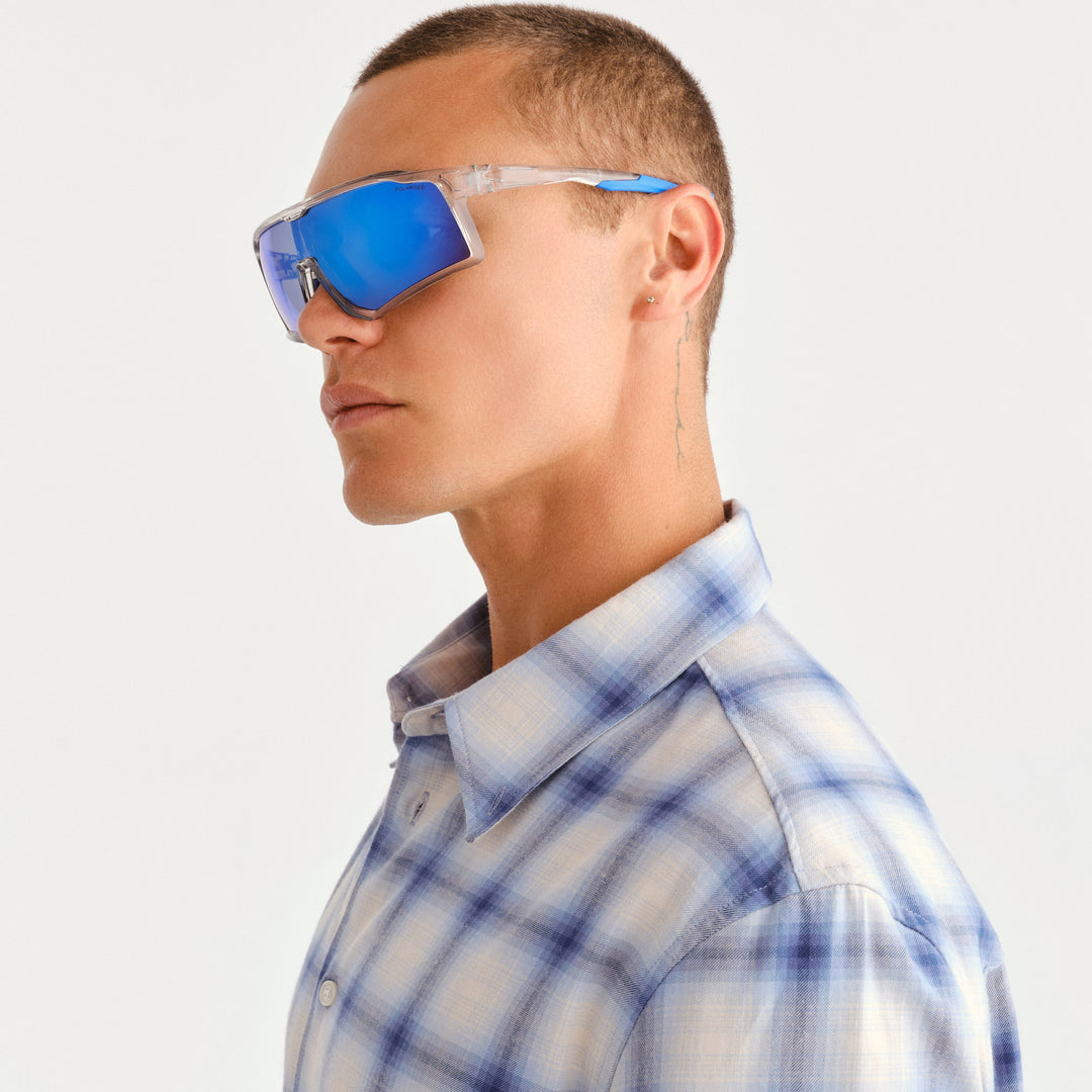 A man in profile wearing oversized shield-style sunglasses with reflective blue lenses and a white frame, paired with a light blue and white plaid button-up shirt against a plain light background.