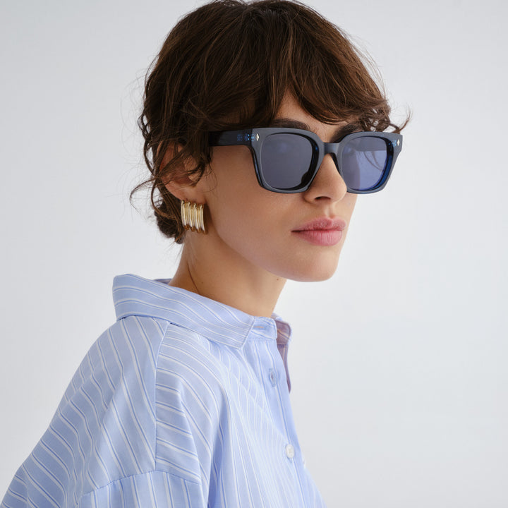 Woman wearing Kingscliff Navy sunglasses and a light blue striped shirt against a white background
