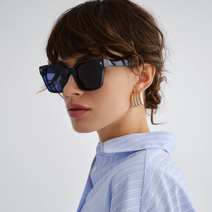 Woman wearing large navy sunglasses and gold hoop earrings on a light gray background