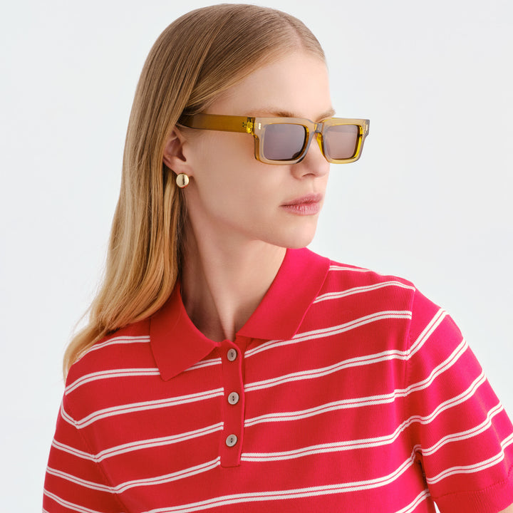 A woman with blonde hair wearing translucent amber square Austinmer sunglasses and a red polo shirt with thin white horizontal stripes, photographed against a clean light gray studio background.