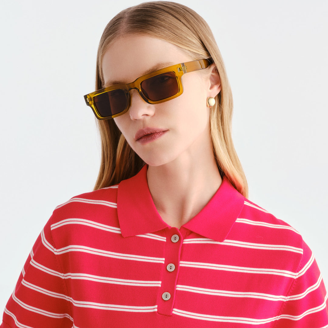 A woman wearing rectangular Austinmer sunglasses with translucent amber-yellow frames and dark lenses, paired with a bright pink and white striped polo shirt, against a clean light gray background.
