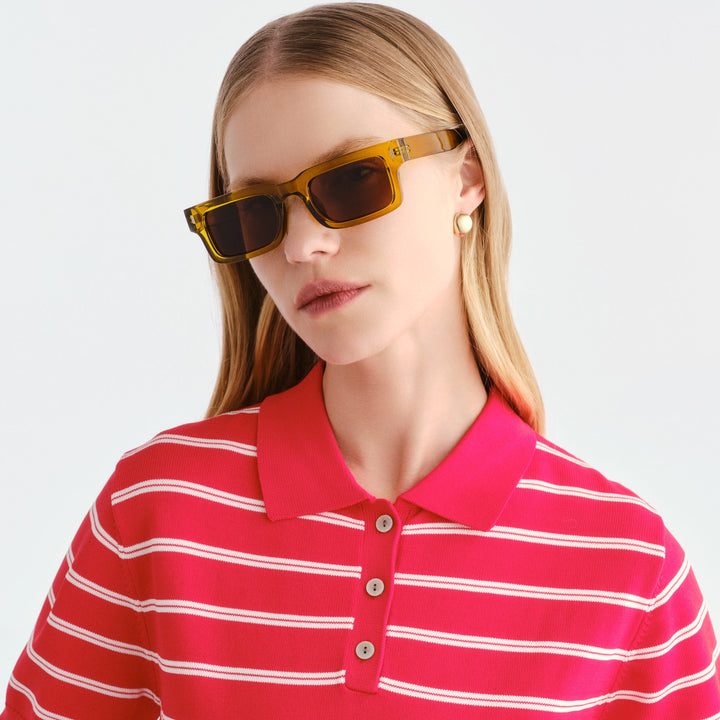 A woman wearing rectangular Austinmer sunglasses with translucent amber-yellow frames and dark lenses, paired with a bright pink and white striped polo shirt, against a clean light gray background.