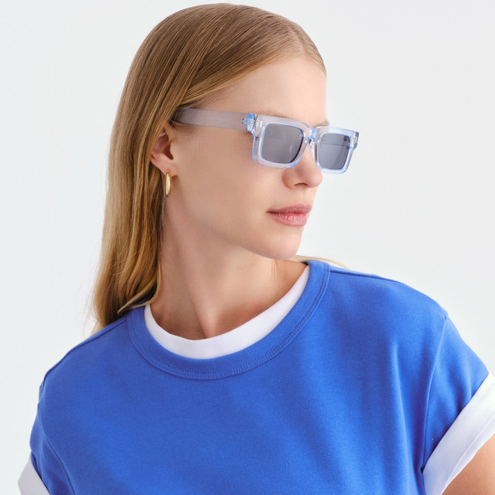 A woman with long light brown hair is shown in profile wearing square Austinmer sunglasses with translucent light blue frames and blue lenses
