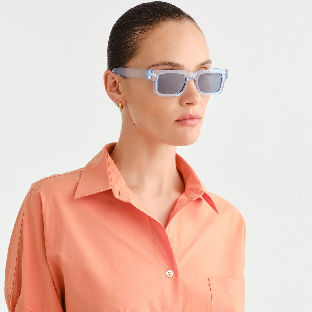 A model with dark hair pulled back wears light blue translucent square sunglasses with dark lenses, paired with a coral-orange button-down shirt, against a light gray studio background.