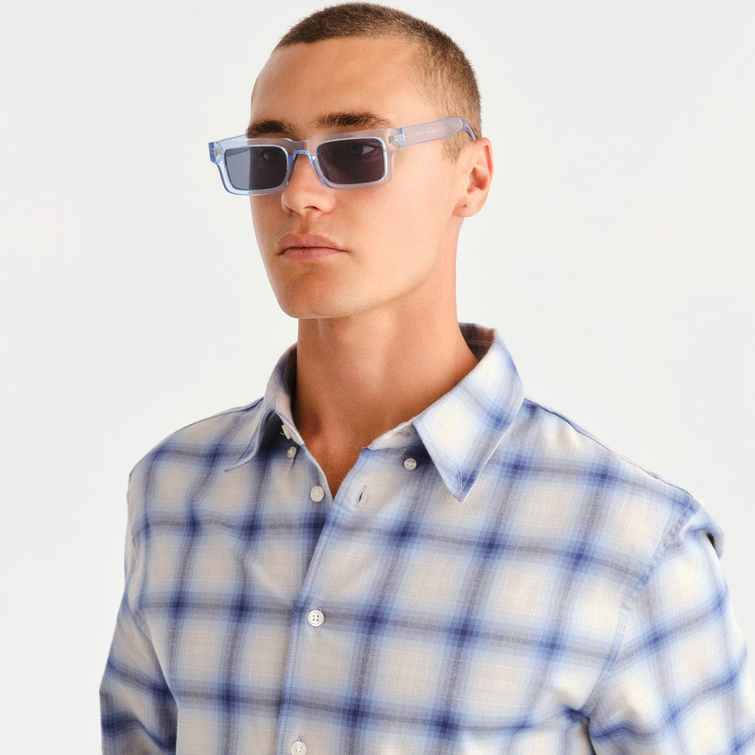 A man wearing light blue translucent square sunglasses with dark lenses and a blue and white plaid button-down shirt, photographed against a clean white background.