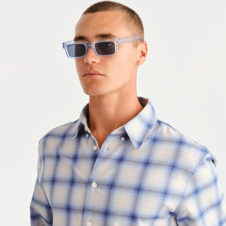 A man wearing light blue translucent square sunglasses with dark lenses and a blue and white plaid button-down shirt, photographed against a clean white background.