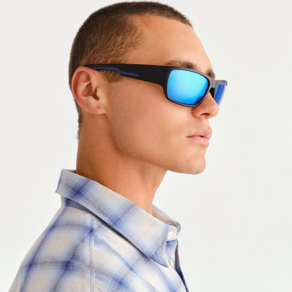 A man in profile wearing black square sunglasses with bright blue mirrored lenses and a blue-and-white plaid button-up shirt, photographed against a clean light gray background.