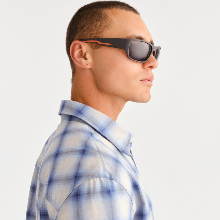 A man in profile wearing dark sunglasses with orange accents and a blue and white plaid button-down shirt, photographed against a clean light gray background.