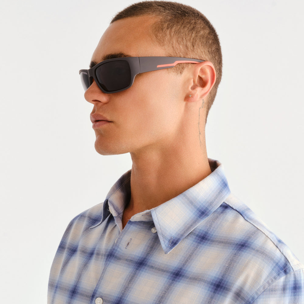 A man in profile wearing dark gray sunglasses with a thin coral-pink accent on the temple arm, paired with a light blue and white plaid button-down shirt against a clean white background.