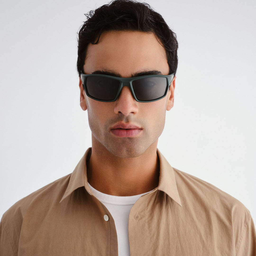 Man wearing dark sunglasses and a beige shirt against a white background