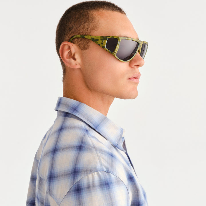 A man in profile wearing rectangular sunglasses with a green camouflage pattern on the frames and a blue and white plaid button-down shirt, set against a plain light gray background.