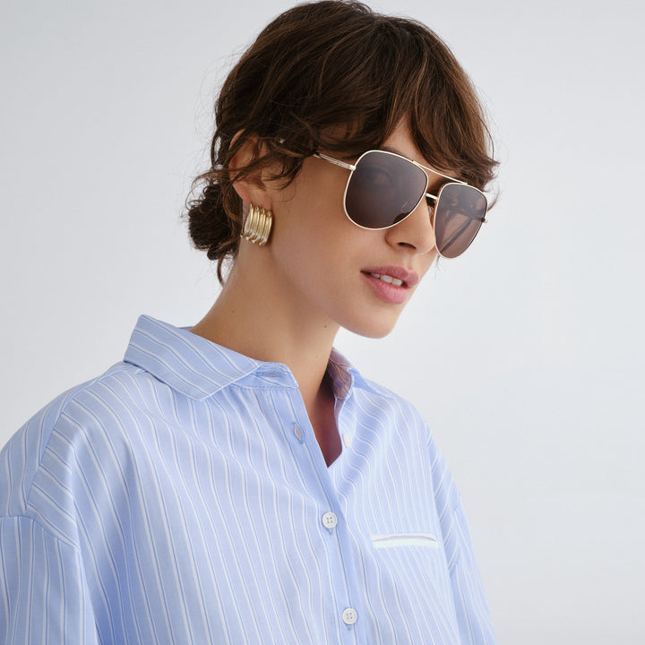 Woman wearing sunglasses and a light blue striped shirt against a plain background